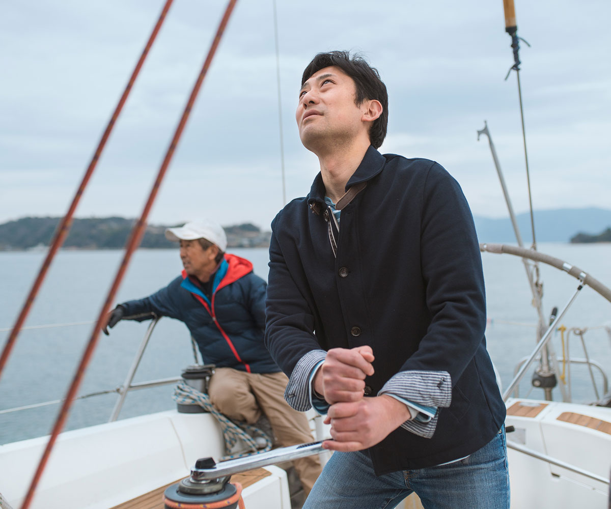 Father and son together on a luxury yacht. The son is hoisting the sail whilst the father relaxes. Okayama, Japan. 